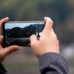 Händer håller en telefon horisontellt, med fingrar som trycker på skärmen för att ta en bild av ett höstlandskap med iPhone-kameran.
