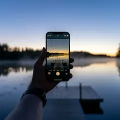 En hand håller upp en iPhone för att ta en bild av en brygga vid en sjö i solnedgången. Perfekt för att redigera bilder på iPhone.