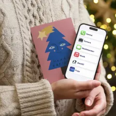 En person i en stickad tröja håller en julkort med en blå gran och en iPhone. På skärmen syns appar som Samtal, Kamera, Meddelanden, Musik, BankID och Freja, vilket antyder ett trepartssamtal via iPhone.