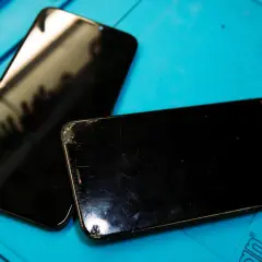 Två svarta mobiltelefoner med spruckna skärmar ligger på ett blått underlag, redo för att laga mobil.
