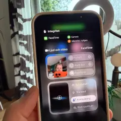 En person håller upp en iPhone med FaceTime-menyn öppen. Kameran visar en person som pratar, med alternativ för ljud och video.