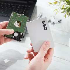 Händer håller upp en traditionell hårddisk och ett modernt SSD-minne bredvid en laptop.