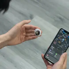 En hand håller en AirTag bredvid en iPhone som visar en karta. Bra airtag batteritid gör den pålitlig.
