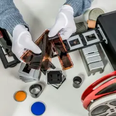 Händer med vita handskar sorterar filmremsor och diabilder för att omvandla negativ till digitala bilder. En filmscanner och en filmrulle syns också.