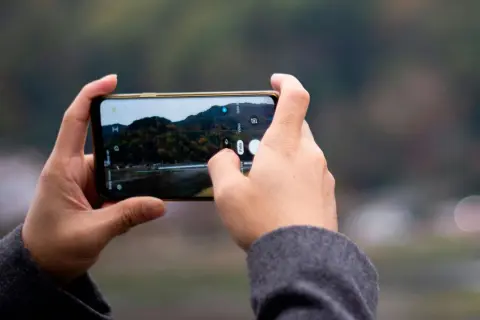 Händer håller en telefon horisontellt, med fingrar som trycker på skärmen för att ta en bild av ett höstlandskap med iPhone-kameran.