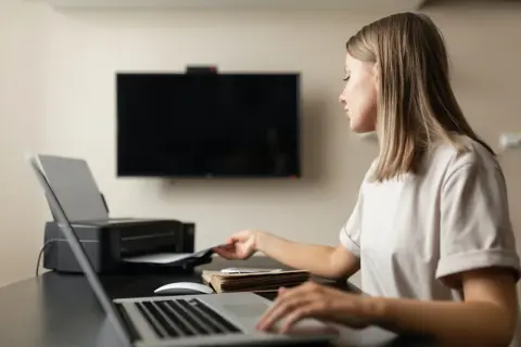 Kvinna vid skrivbord med laptop och skrivare. Hon tar papper ur skrivaren, redo att skriva ut i A4 storlek pixlar.