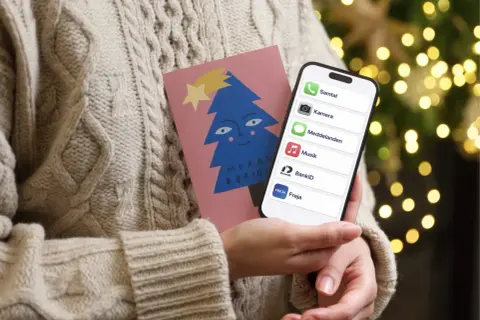 En person i en stickad tröja håller en julkort med en blå gran och en iPhone. På skärmen syns appar som Samtal, Kamera, Meddelanden, Musik, BankID och Freja, vilket antyder ett trepartssamtal via iPhone.