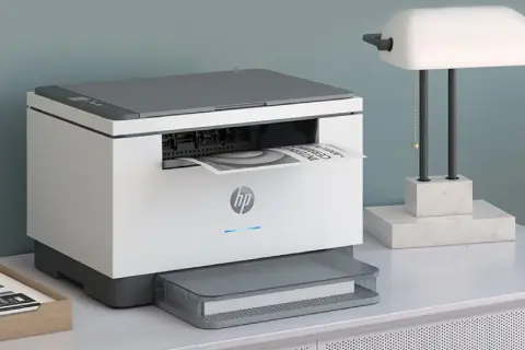 En vit och grå HP-skrivare, den minsta laserskrivaren, skriver ut ett dokument. Bredvid står en lampa med marmorbas.