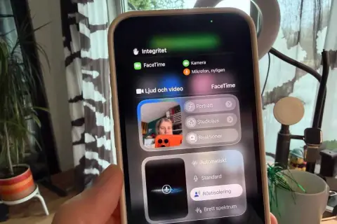 En person håller upp en iPhone med FaceTime-menyn öppen. Kameran visar en person som pratar, med alternativ för ljud och video.