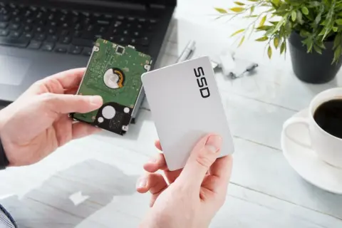 Händer håller upp en traditionell hårddisk och ett modernt SSD-minne bredvid en laptop.