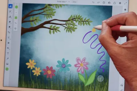 Handen skapar en magisk virvel med en penna i Adobe Fresco, över en bild av en drömsk äng med blommor och träd.