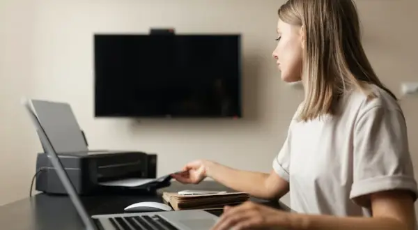 Kvinna vid skrivbord med laptop och skrivare. Hon tar papper ur skrivaren, redo att skriva ut i A4 storlek pixlar.