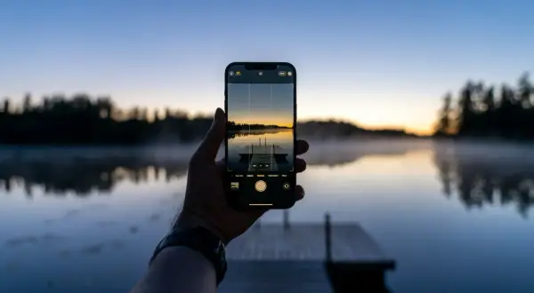 En hand håller upp en iPhone för att ta en bild av en brygga vid en sjö i solnedgången. Perfekt för att redigera bilder på iPhone.