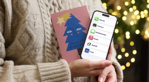 En person i en stickad tröja håller en julkort med en blå gran och en iPhone. På skärmen syns appar som Samtal, Kamera, Meddelanden, Musik, BankID och Freja, vilket antyder ett trepartssamtal via iPhone.