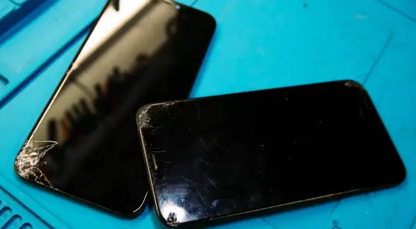 Två svarta mobiltelefoner med spruckna skärmar ligger på ett blått underlag, redo för att laga mobil.