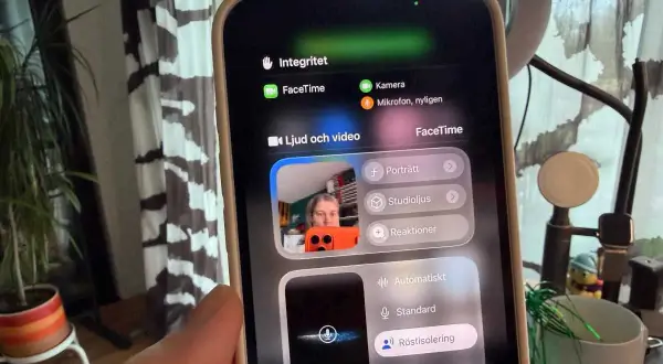 En person håller upp en iPhone med FaceTime-menyn öppen. Kameran visar en person som pratar, med alternativ för ljud och video.