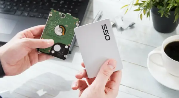 Händer håller upp en traditionell hårddisk och ett modernt SSD-minne bredvid en laptop.