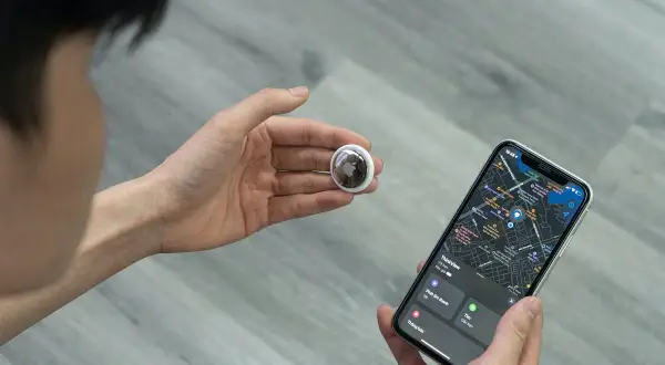 En hand håller en AirTag bredvid en iPhone som visar en karta. Bra airtag batteritid gör den pålitlig.