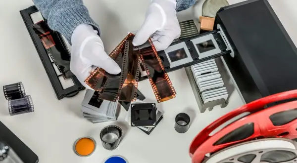 Händer med vita handskar sorterar filmremsor och diabilder för att omvandla negativ till digitala bilder. En filmscanner och en filmrulle syns också.