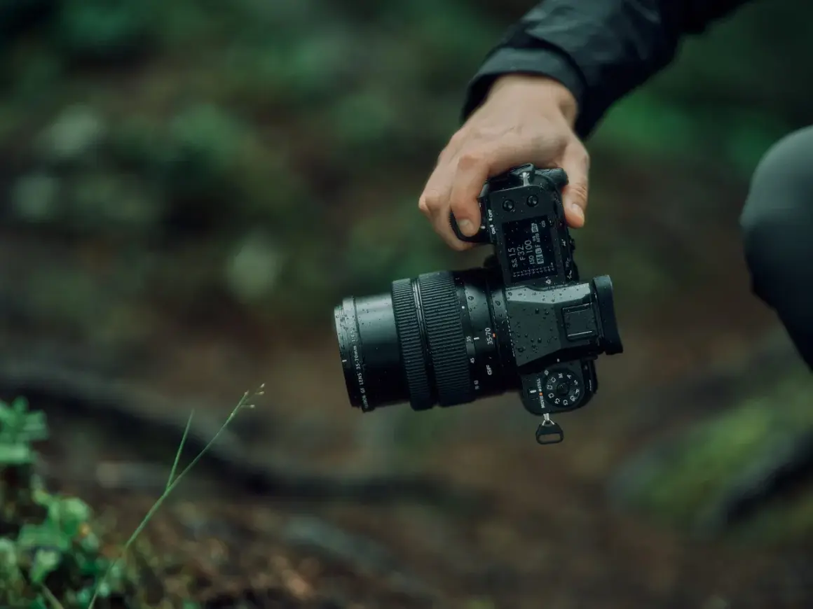 En hand håller en Fujifilm GFX 50S II med ett objektiv i en regnig skogsmiljö.