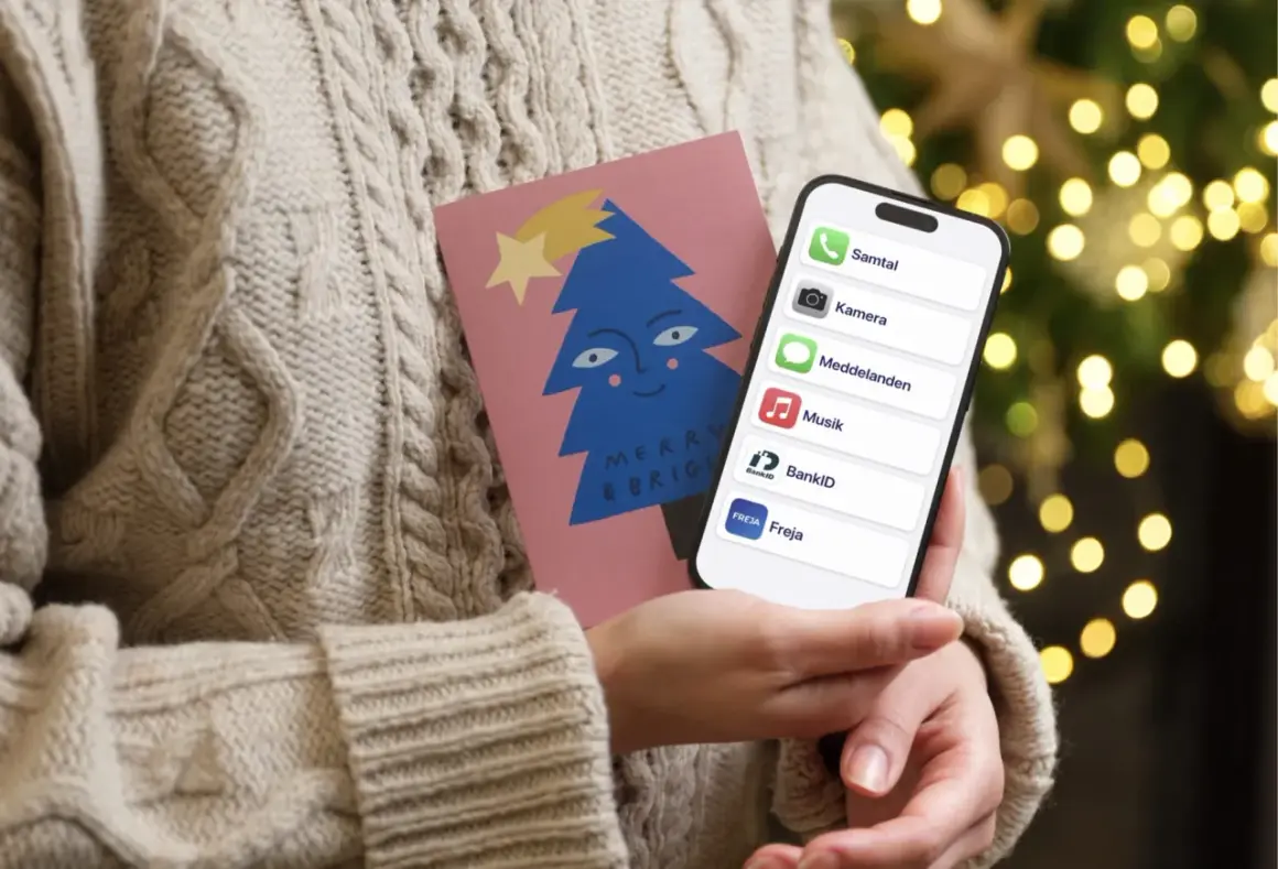 En person i en stickad tröja håller en julkort med en blå gran och en iPhone. På skärmen syns appar som Samtal, Kamera, Meddelanden, Musik, BankID och Freja, vilket antyder ett trepartssamtal via iPhone.