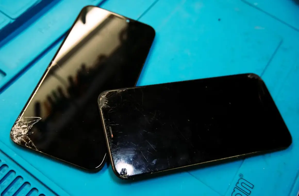 Två svarta mobiltelefoner med spruckna skärmar ligger på ett blått underlag, redo för att laga mobil.