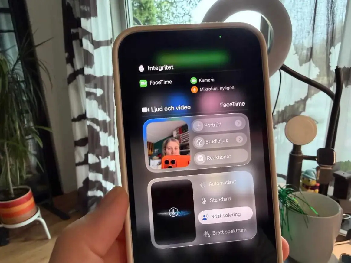 En person håller upp en iPhone med FaceTime-menyn öppen. Kameran visar en person som pratar, med alternativ för ljud och video.