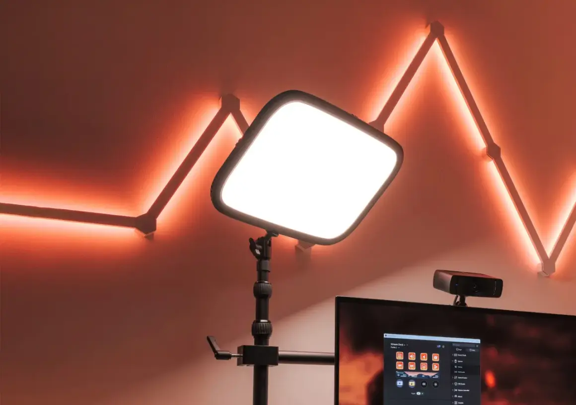 En stor, ljusstark key light lyser upp scenen. Bakom den syns en datorskärm med Stream Deck-mjukvara och en webbkamera. Väggen pryds av en geometrisk ljusslinga i orange.
