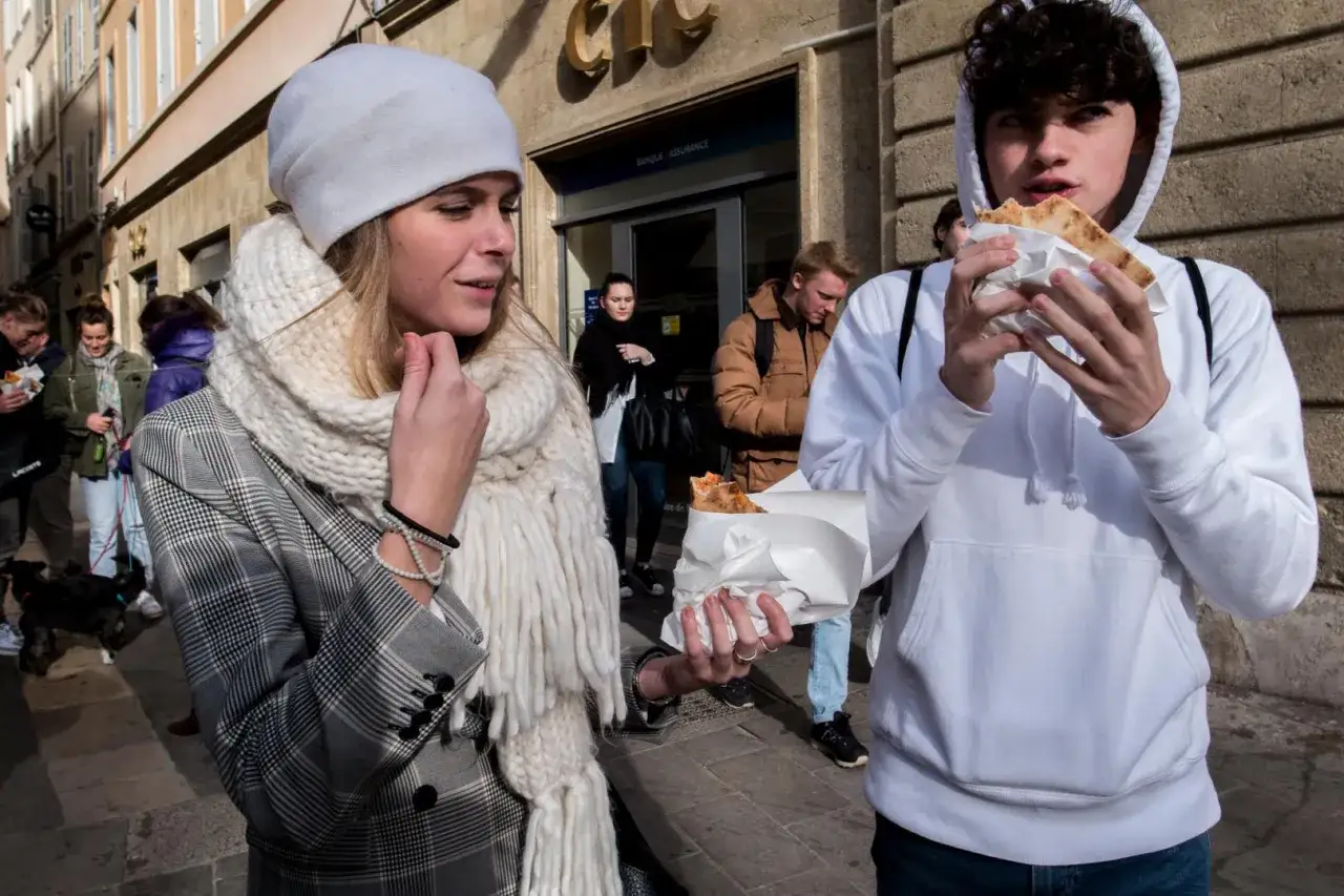 En ung kvinna med vit mössa och stor halsduk tittar på en kille som äter pizza. En scen fångad med en Ricoh GR IV.