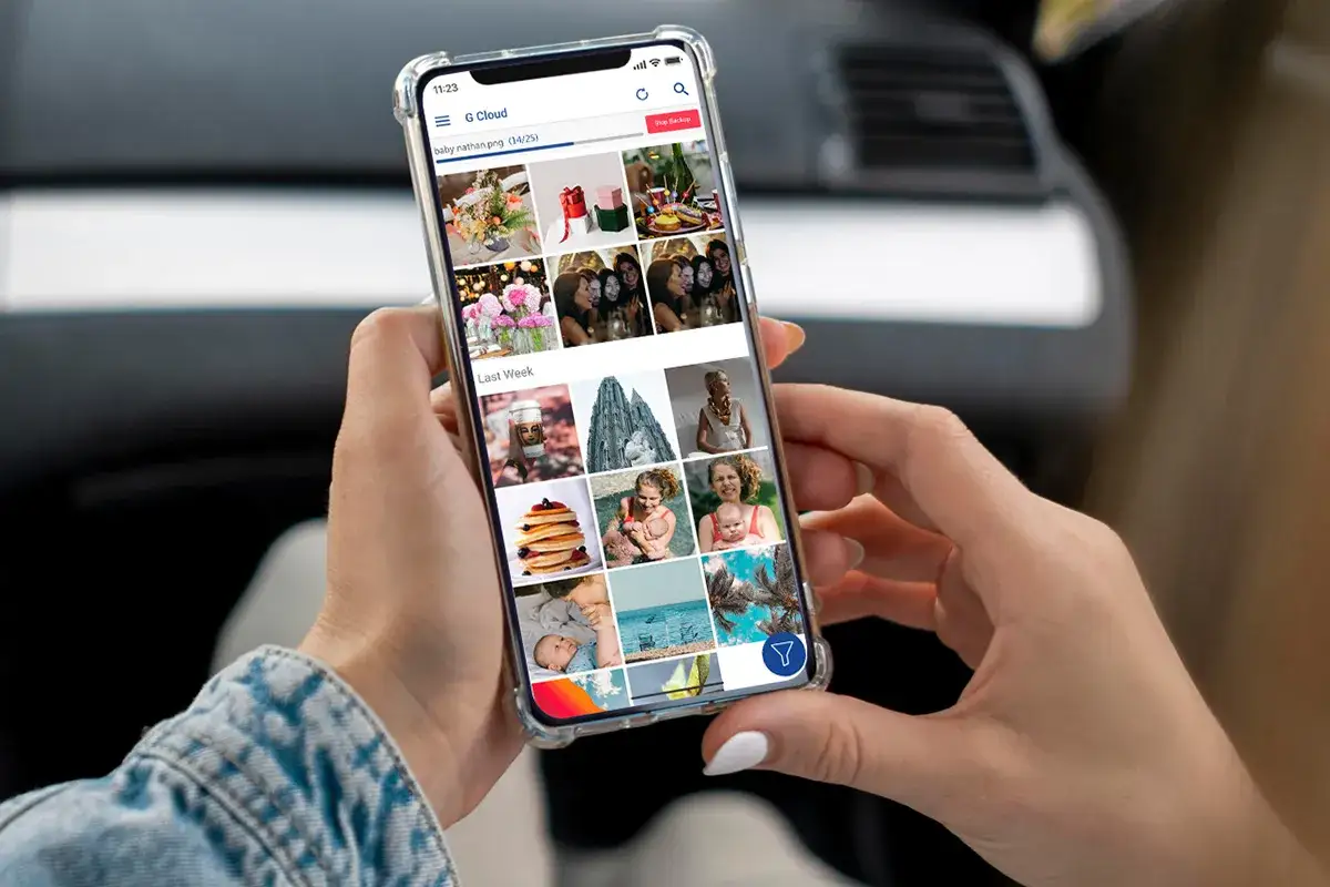 En person h&aring;ller en mobil som visar G Cloud-appen med bilder. Perfekt f&ouml;r att spara bilder fr&aring;n mobilen och organisera minnen.