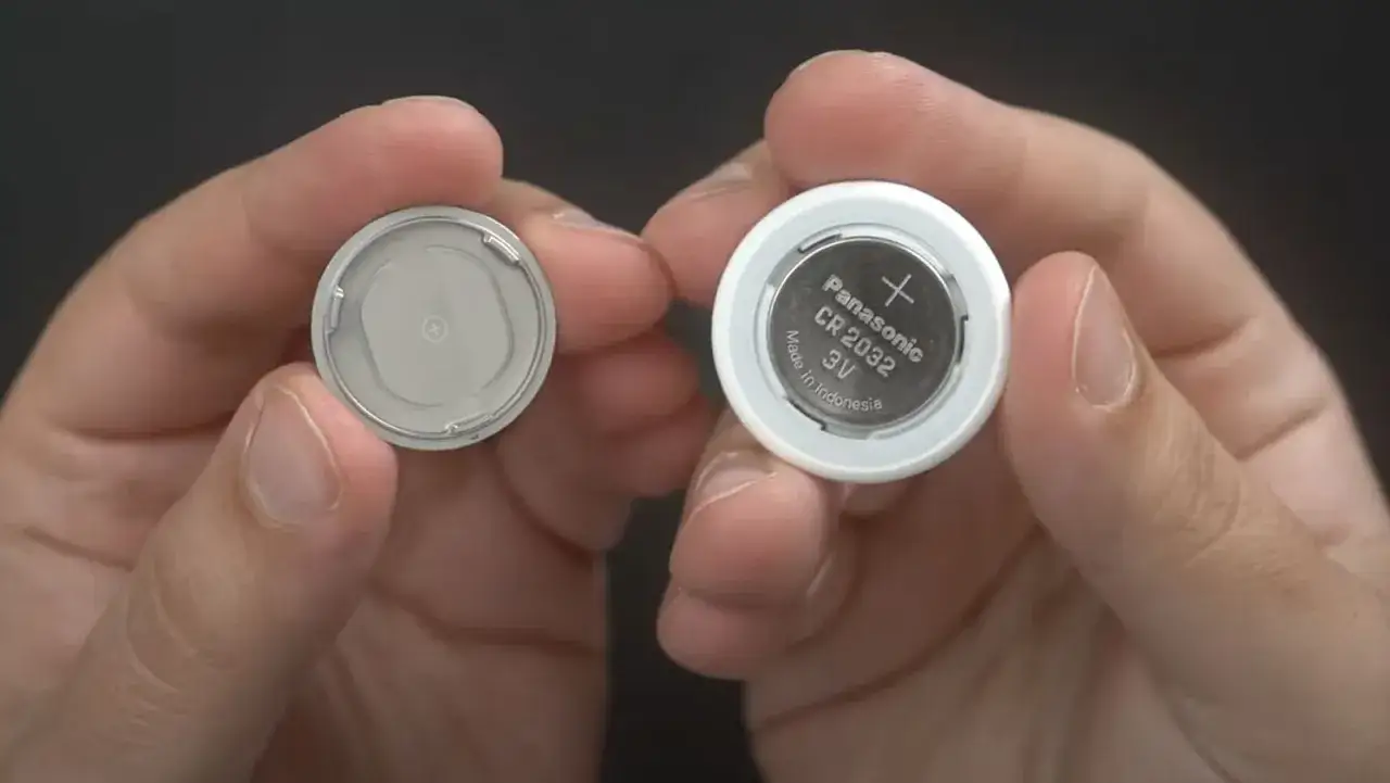 Två händer håller upp en AirTag. Till höger syns en vit AirTag med ett Panasonic CR 2032 batteri. Till vänster syns det avtagna locket. Bra airtag batteritid!