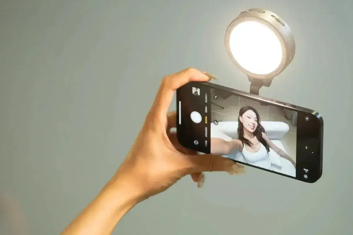 En hand håller en mobiltelefon för att ta en selfie bild. En ringlampa lyser upp ansiktet.
