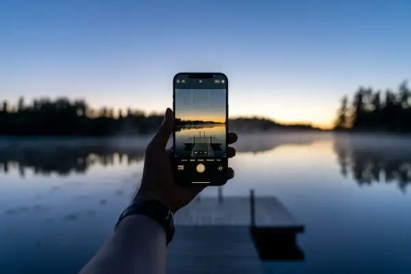 En hand håller upp en iPhone för att ta en bild av en brygga vid en sjö i solnedgången. Perfekt för att redigera bilder på iPhone.