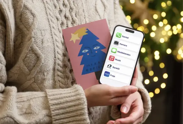 En person i en stickad tröja håller en julkort med en blå gran och en iPhone. På skärmen syns appar som Samtal, Kamera, Meddelanden, Musik, BankID och Freja, vilket antyder ett trepartssamtal via iPhone.