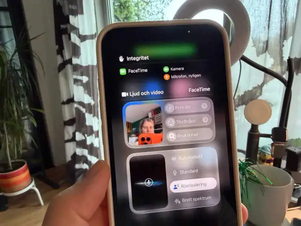 En person håller upp en iPhone med FaceTime-menyn öppen. Kameran visar en person som pratar, med alternativ för ljud och video.